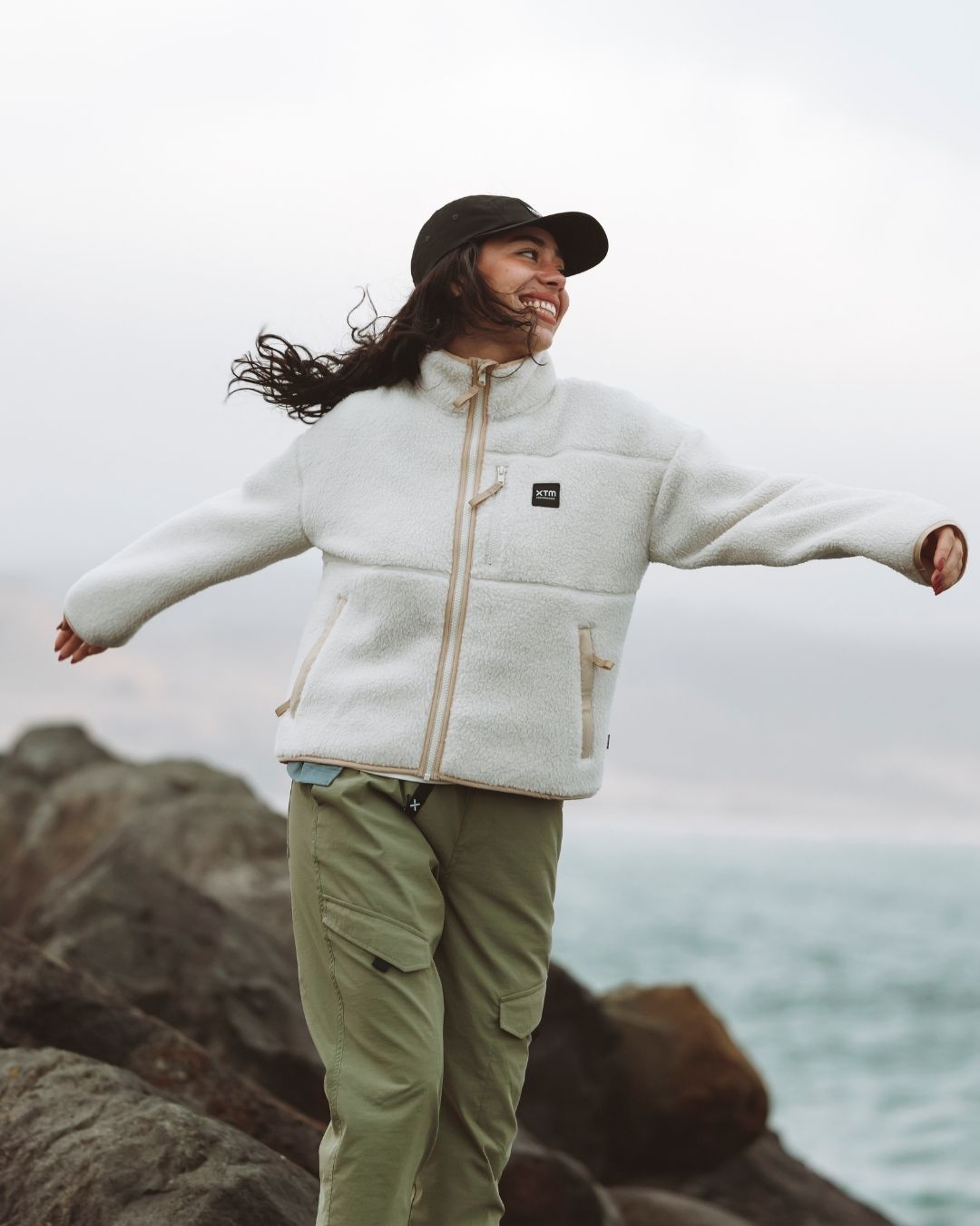 Woman in a cream fleece with arms out on coastal rocks, wearing the black XTM Hakea cap