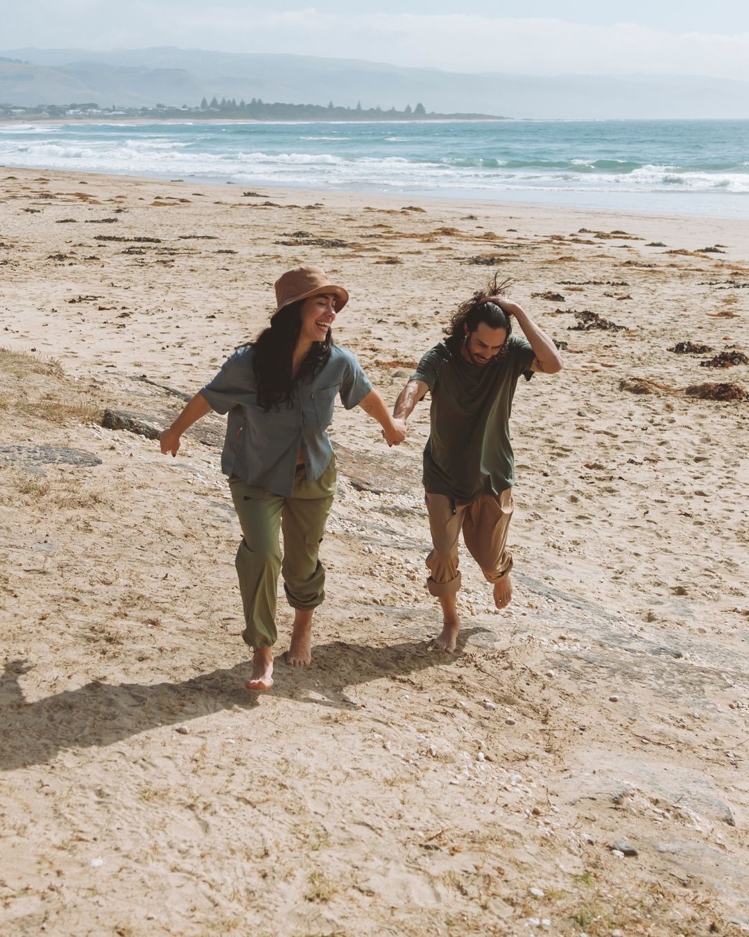Couple running barefoot along a beach, the woman in the Woodsmoke XTM Sundew bucket hat