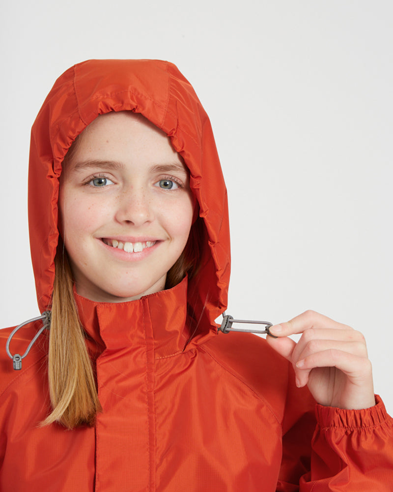 Smiling girl in the XTM Kids Stash 2L rain jacket in Burnt Orange, hood up and toggle pulled