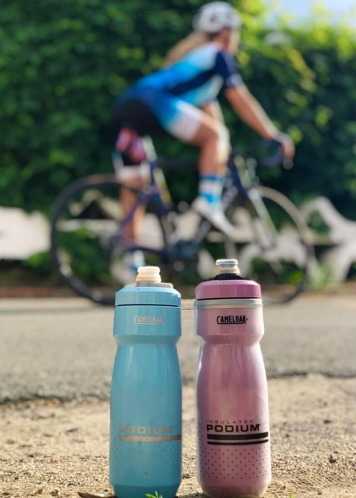 Blue and purple CamelBak Podium bottles standing on a road with a cyclist riding past behind
