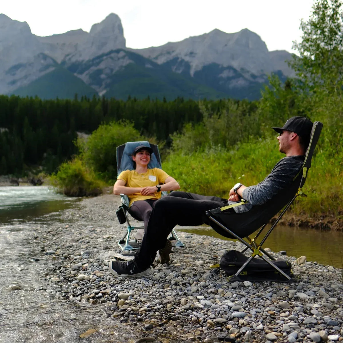Two campers in Nemo chairs on a gravel riverbank below rocky mountains