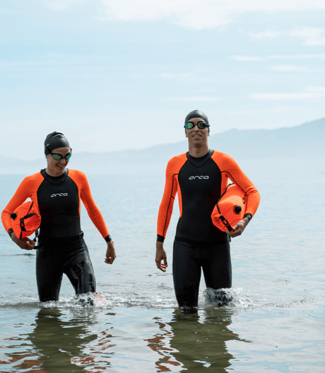 Two swimmers in Orca wetsuits wading out of the sea carrying orange tow floats