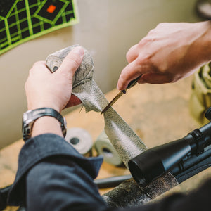 Hands cutting a strip of Camo Form with a knife beside a scoped rifle