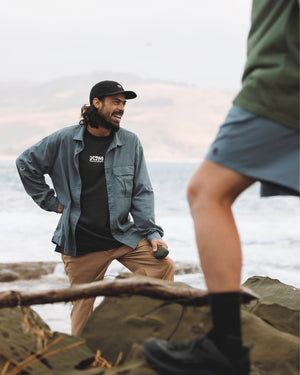 Man in a denim shirt on the rocks by the sea, wearing the black XTM Hakea cap