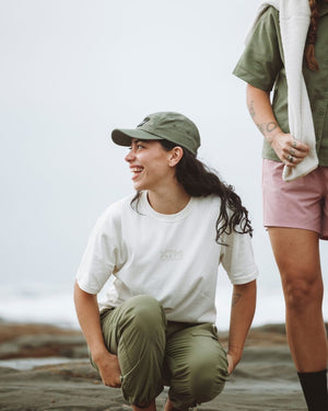 Woman crouching on a rocky shoreline wearing the XTM Hakea cap in Thyme with a white tee
