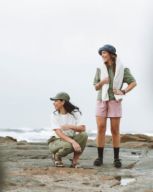 Two women on a coastal rock shelf, one standing in the Indigo XTM Sundew bucket hat