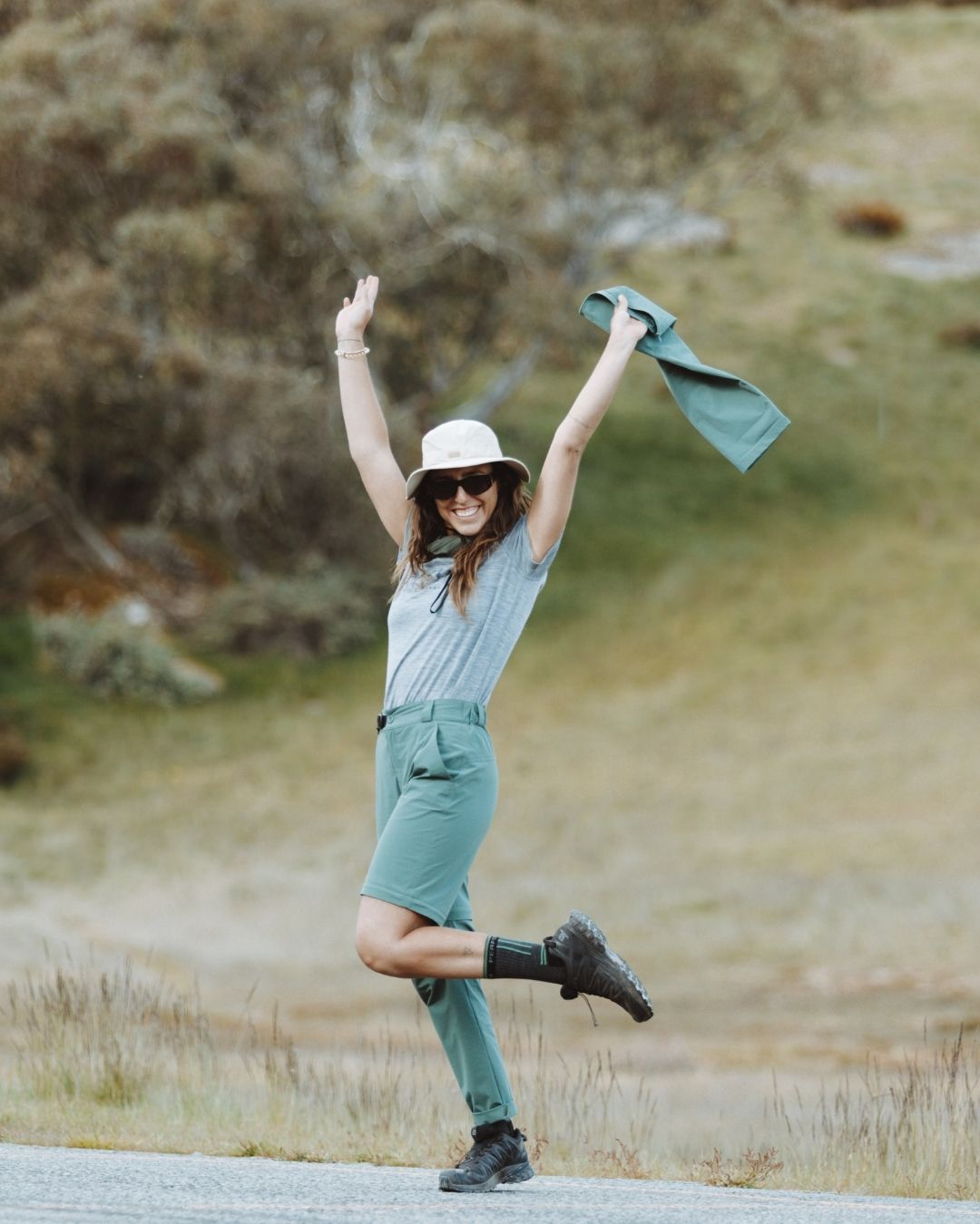 Woman leaping on a grassy hillside in XTM Wye River Spruce Green hiking pants and sun hat