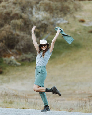 Woman leaping on a grassy hillside in XTM Wye River Spruce Green hiking pants and sun hat