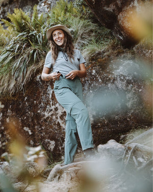 Smiling woman in sun hat beside a mossy rock wearing XTM Wye River Spruce Green pants