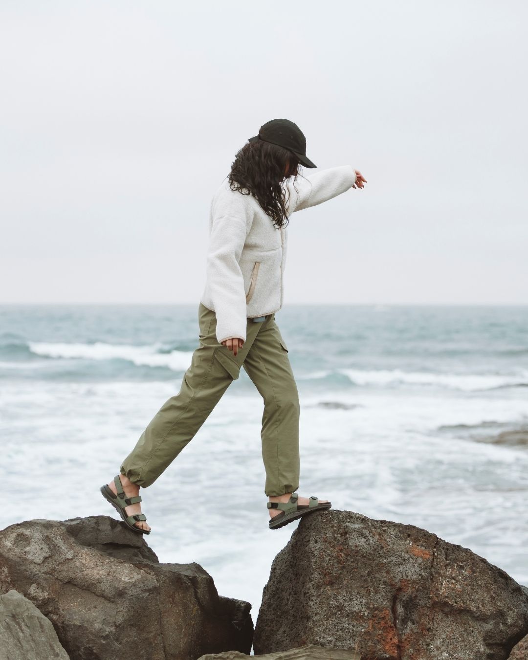 Woman in XTM Acacia Olive hiking pants and sandals striding between rocks by the sea
