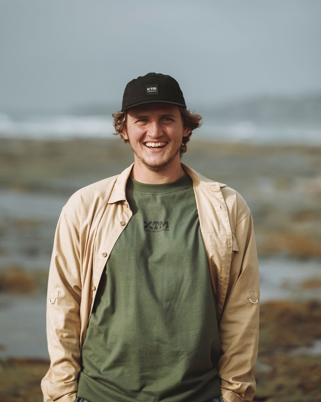 Smiling man wearing the Dune XTM Messmate shirt open over a green tee by the sea
