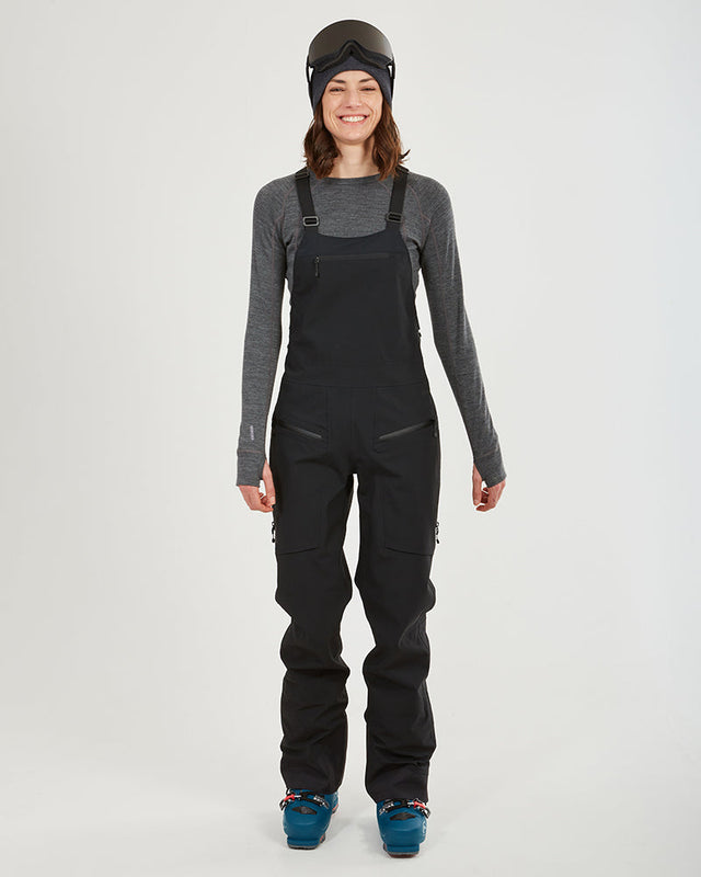 Woman wearing the black XTM Stellar 2.0 hardshell bib pant with ski boots, front view