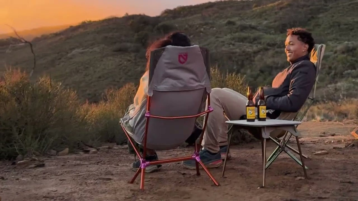 Two people relaxing in camp chairs on a scrubby hillside at sunset