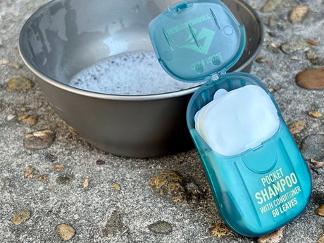 Open teal Pocket Shampoo case showing the soap leaves, beside a bowl of suds on rock